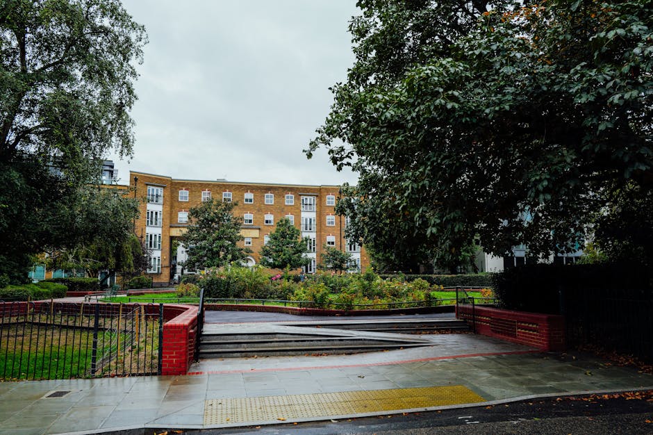 A well-maintained public garden in Woolwich with a symmetrical layout featuring two paved pathways flanking a central flower bed filled with low-growing, flowering plants and green foliage. The pathwa
